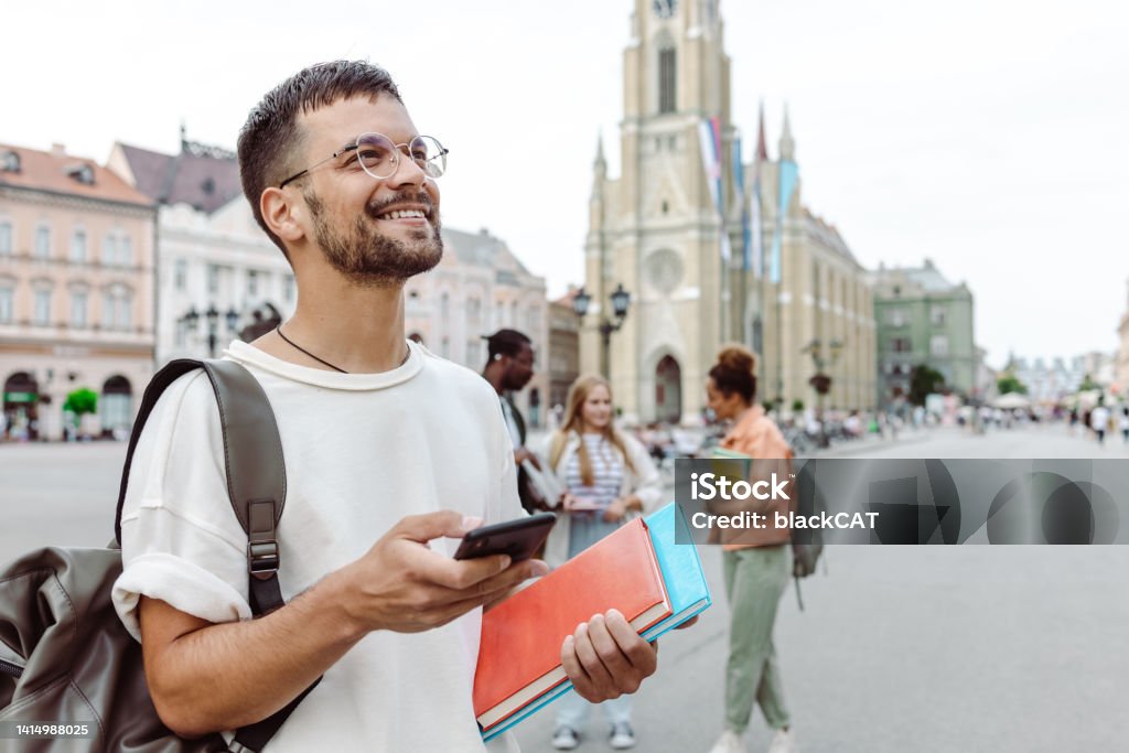 istockphoto-1414988025-1024×1024 A young man carrying notebooks and using smart phone on the street while walking. A group of other students is behind